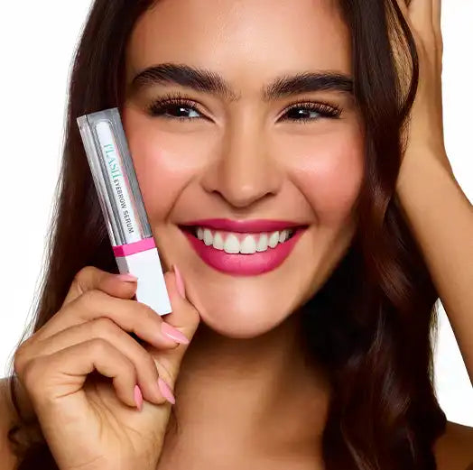 Smiling woman holding fLASH Brow Serum tube, showcasing full brows and natural makeup