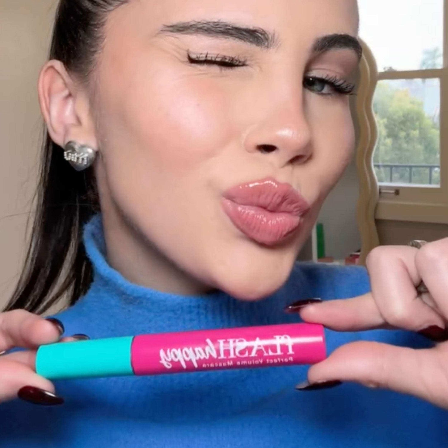 Woman with full lashes holding fLASH Cosmetics mascara, wearing blue sweater, closeup